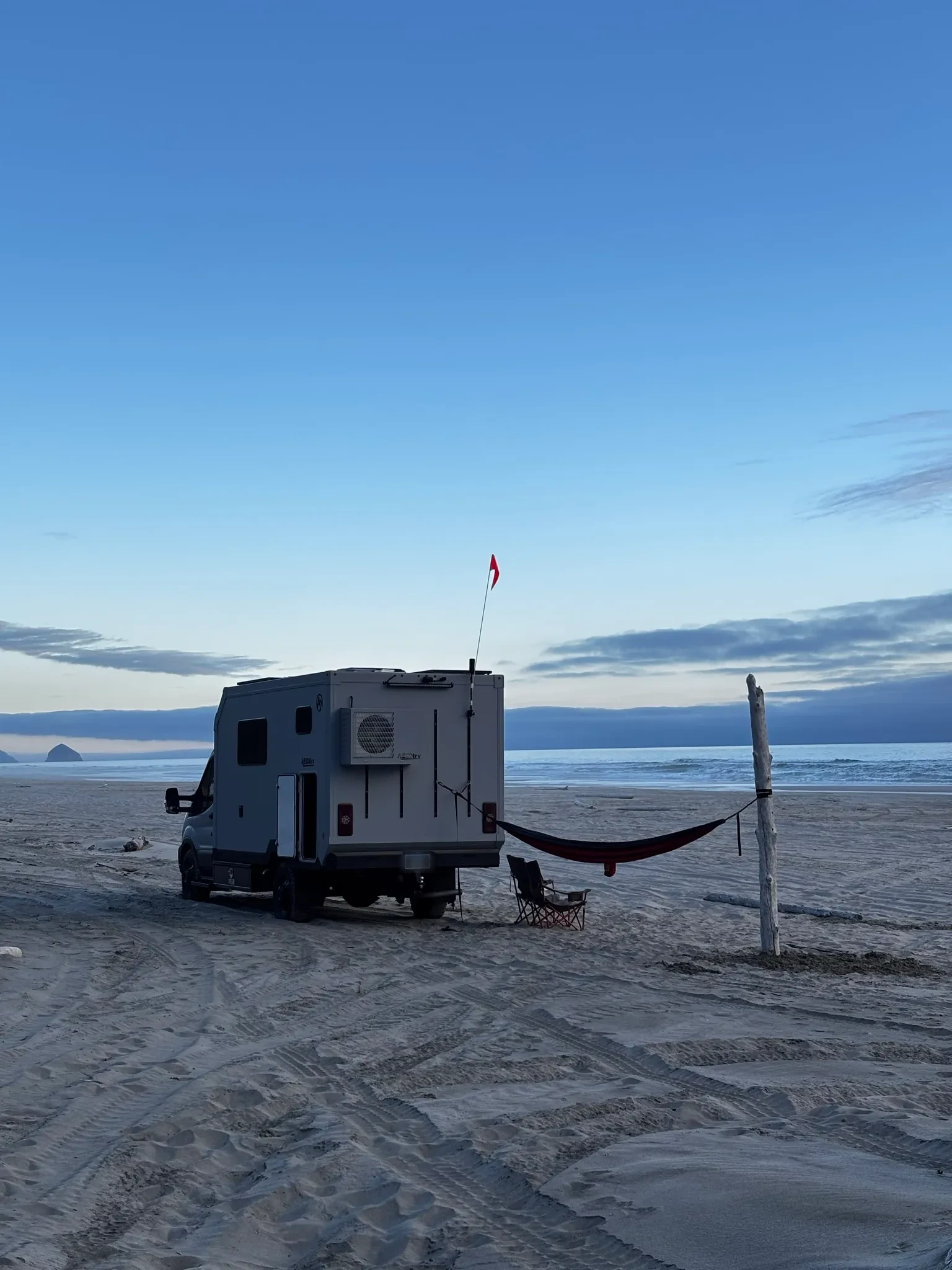 A white AEONrv coach on a Transit cutaway chassis parked on wet sand at dusk, with a roof mounted air conditioner on the rear wall, a pennant flag on an antenna, and a hammock strung between the rig and a driftwood post