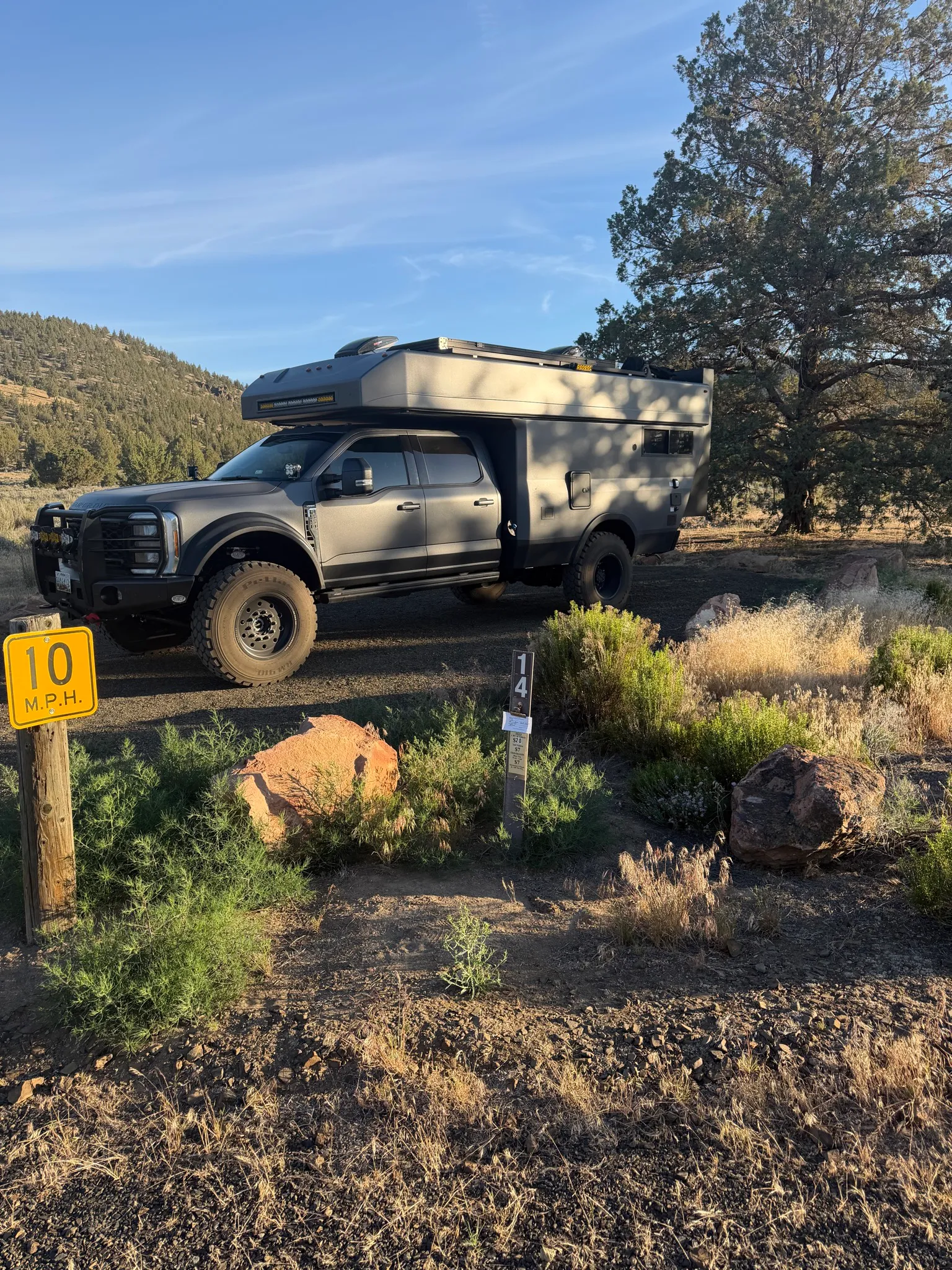 A grey Ford Super Duty crew cab carrying a flatbed pop-top truck camper, parked at a gravel campsite among juniper trees, with oversized off-road tires, a front brush guard, and a spare tire mounted on the camper's side